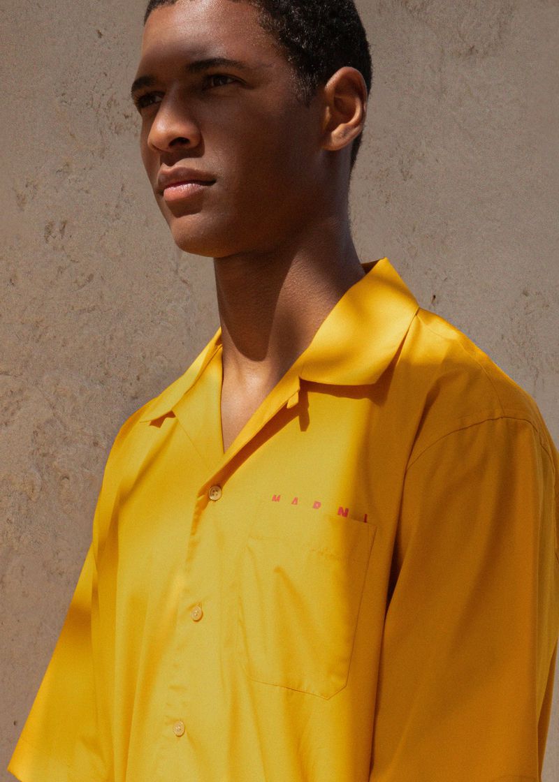 A portrait of a handsome adult male in a vibrant yellow shirt against a neutral wall, bathed in soft, diffused sunlight, showcasing fashion apparel.