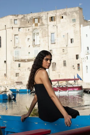 Adult female poses in a black dress, seated on a boat against a backdrop of a weathered building and blue sky in a harborside setting.