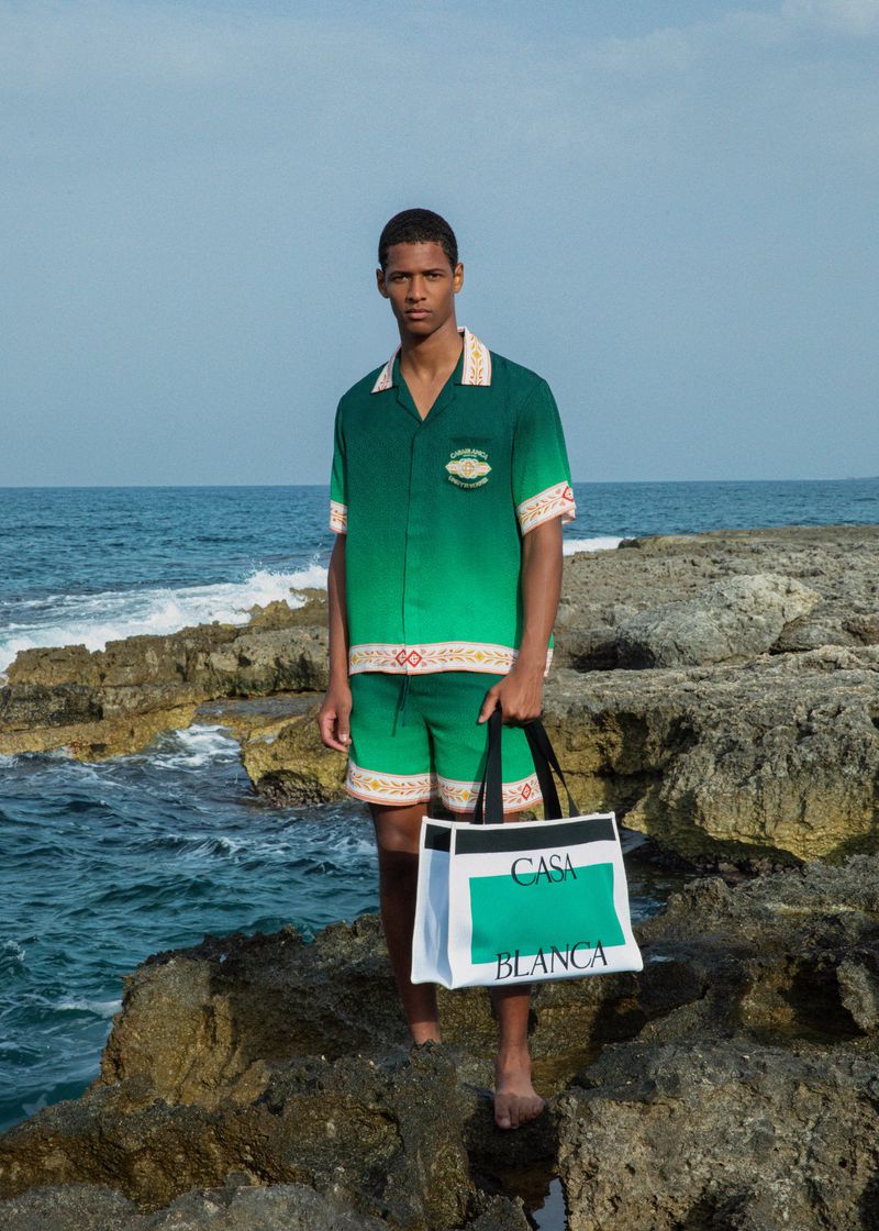 Man in green outfit stands on rocky beach holding bag in fashion shot with ocean, horizon, and blue sky backdrop under daylight.