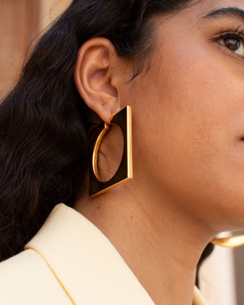 Portrait of an adult female wearing a cream jacket and modern gold geometric earrings, indoor setting, close-up shot.