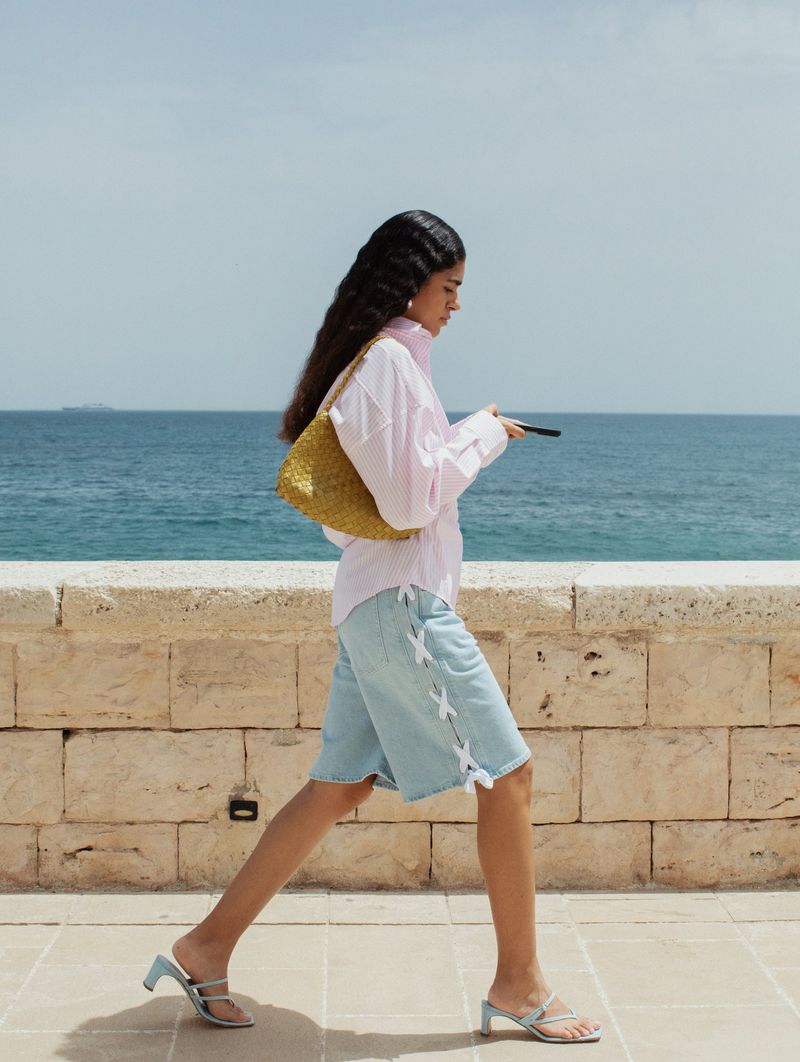 Woman in pink shirt and denim shorts walks along stone wall near blue sea carrying woven purse and holding phone in daylight.