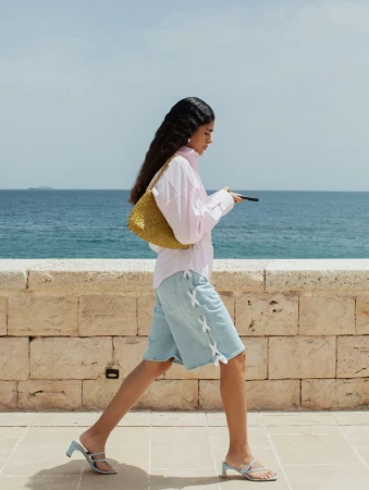 Woman in pink shirt and denim shorts walks along stone wall near blue sea carrying woven purse and holding phone in daylight.