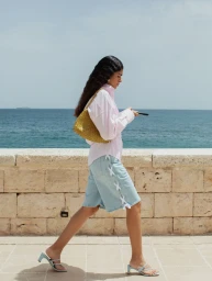 Woman in profile walking along a stone sea wall wearing a striped shirt and denim shorts holding a woven handbag