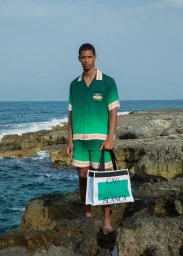 Young man in a green ombre short-sleeve shirt and matching shorts holding a white tote bag on a rocky coast