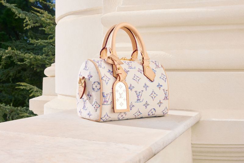 Luxury Louis Vuitton handbag with signature monogram pattern sits outdoors on a bright stone ledge near green foliage, bathed in sunlight.