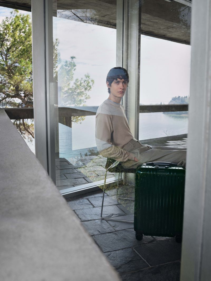 Young man in casual clothes sitting on a chair with a dark green suitcase in a modern architectural space with large windows.