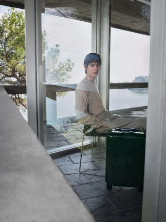 Young man in casual clothes sitting on a chair with a dark green suitcase in a modern architectural space with large windows.