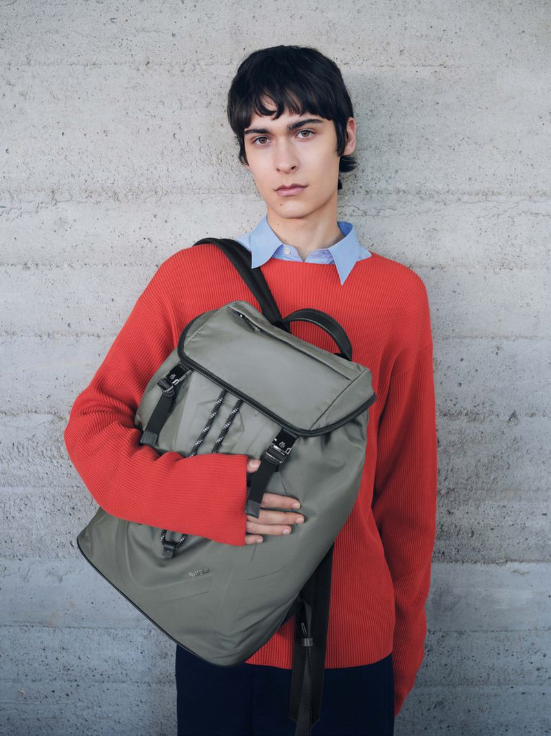 Man in red sweater poses with backpack against neutral wall. Fashion shot focuses on model and accessories. Studio lighting, medium framing.