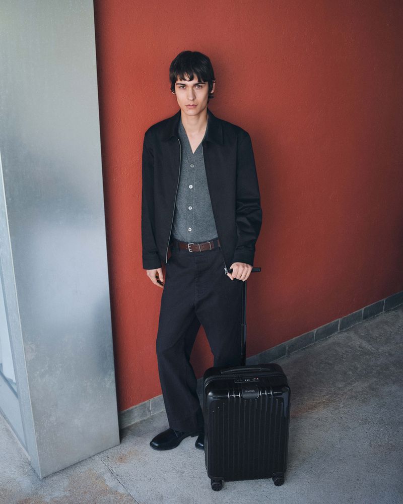 Young man in a jacket and trousers stands with a black suitcase against a brick wall, looking towards the camera in an indoor setting.