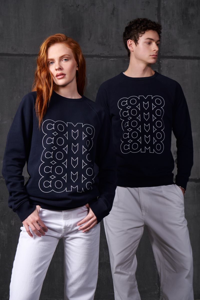 Two people, an adult woman and an adult man, pose indoors wearing matching dark blue sweatshirts and white pants. Fashion shot.