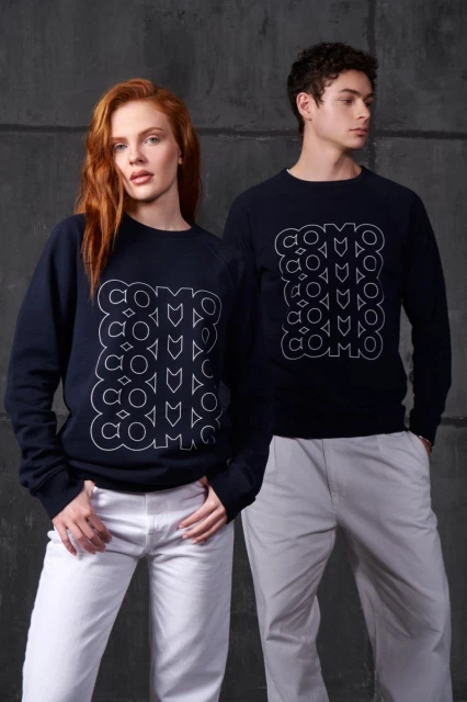 Two people, an adult woman and an adult man, pose indoors wearing matching dark blue sweatshirts and white pants. Fashion shot.