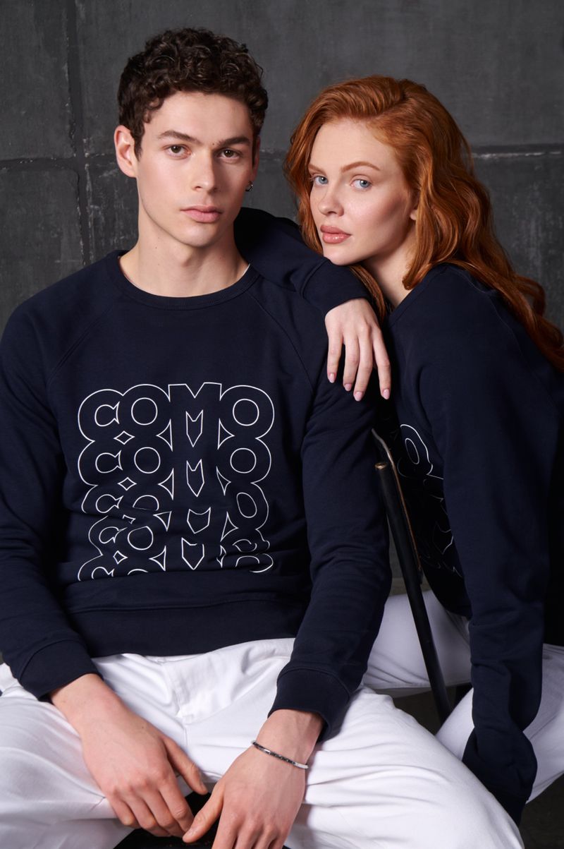 Two fashion models wearing matching navy sweatshirts pose in a photo studio against a gray backdrop. Studio lighting and medium shot.