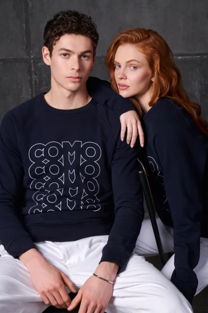 Two fashion models wearing matching navy sweatshirts pose in a photo studio against a gray backdrop. Studio lighting and medium shot.
