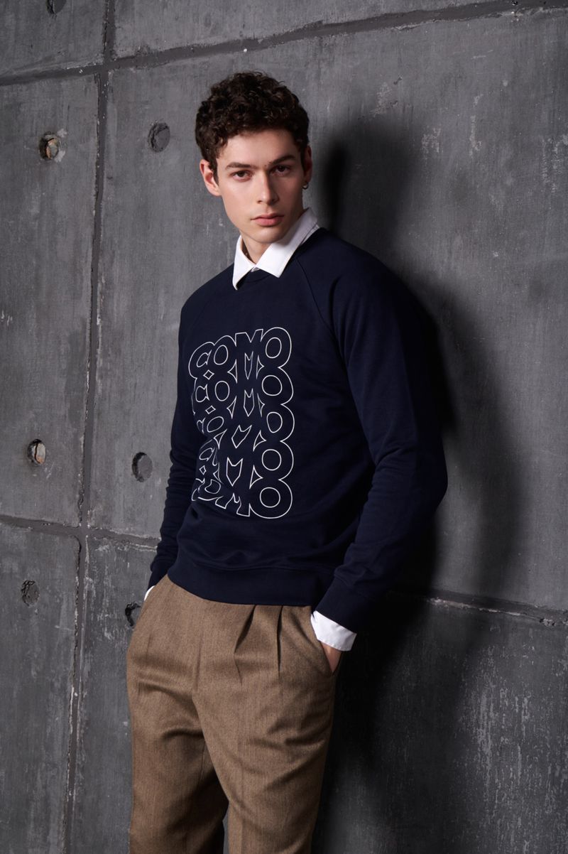 Portrait of a young adult male wearing a navy sweatshirt with a white graphic design and brown trousers in front of a concrete wall.
