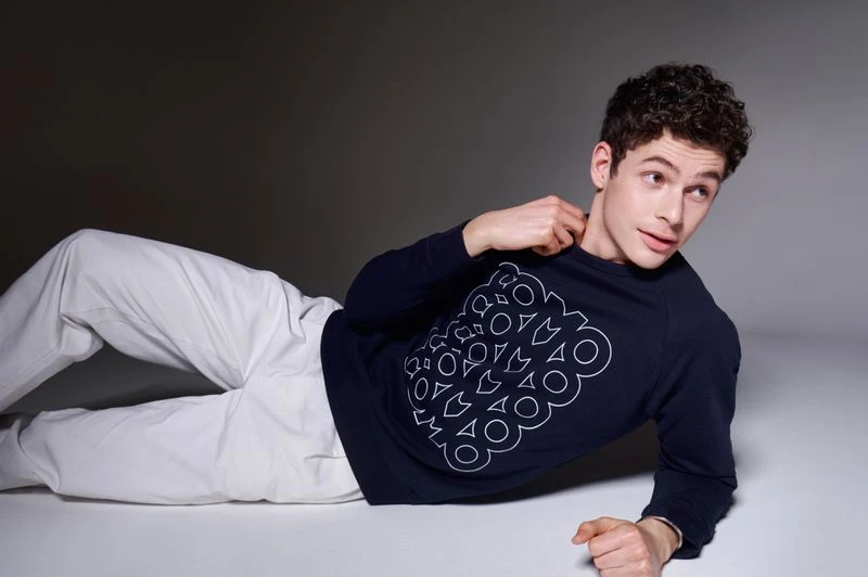 Young adult male poses in a fashion shot, lying on a white surface with a navy patterned sweatshirt and white pants, studio setting.