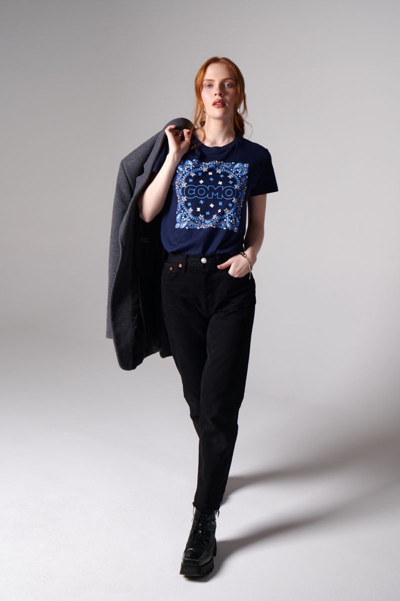 A woman in a t-shirt, jeans, and boots poses in a full shot, holding a grey jacket over her shoulder, set against a light grey background.
