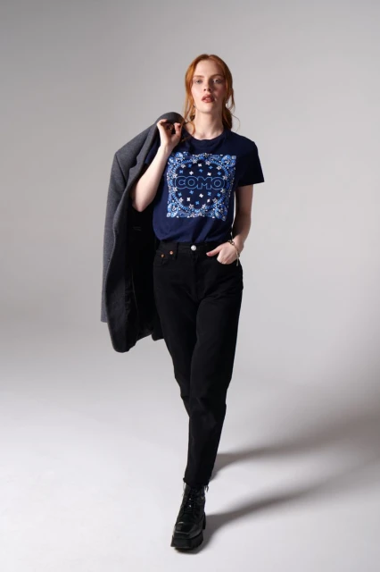 A woman in a t-shirt, jeans, and boots poses in a full shot, holding a grey jacket over her shoulder, set against a light grey background.