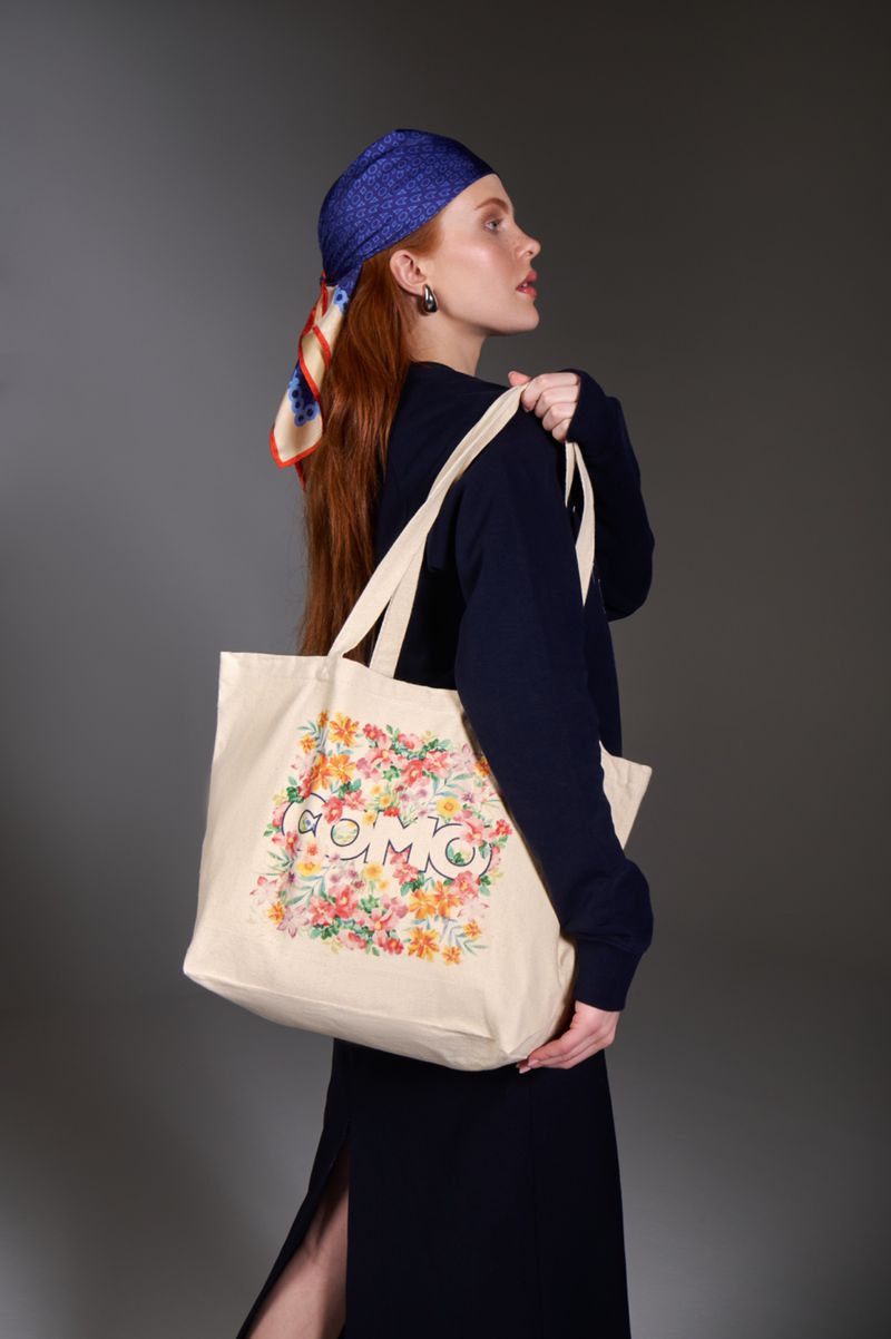 Red-haired woman in a blue headscarf holds a floral tote bag with dark blue dress against a gray background in a studio shot.