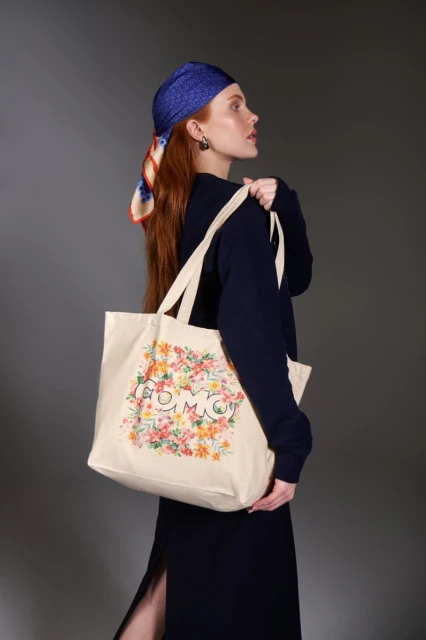 Red-haired woman in a blue headscarf holds a floral tote bag with dark blue dress against a gray background in a studio shot.
