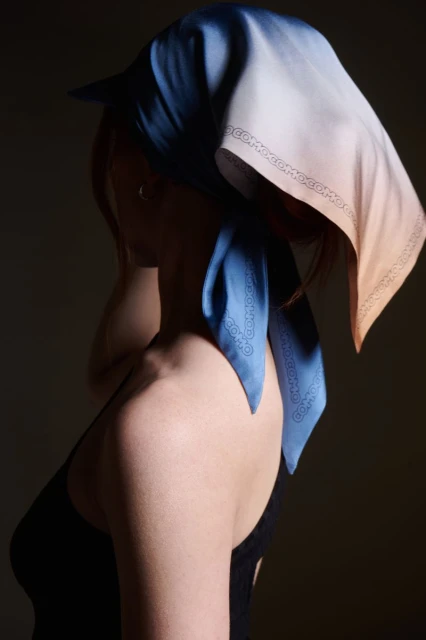 Woman wearing a headscarf with blue and peach tones, standing against a dark background, fashion portrait highlighting the scarf's design.