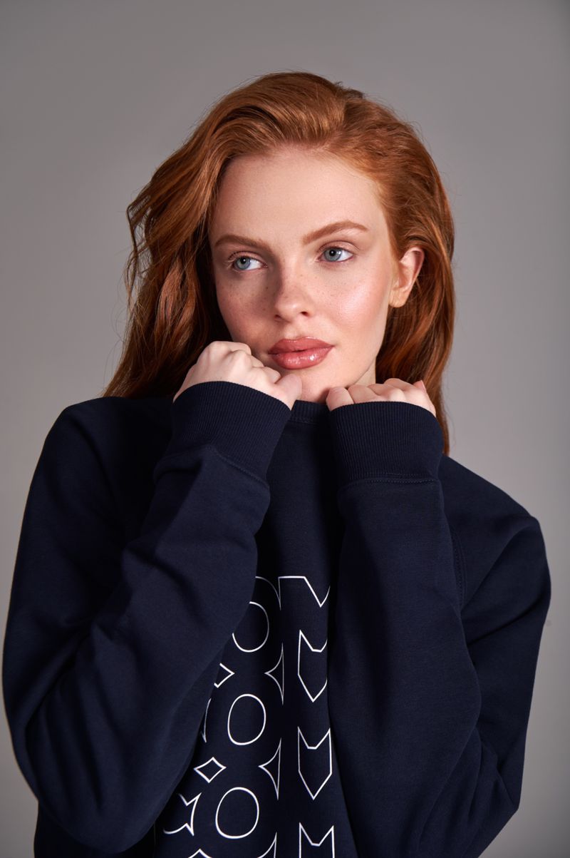 Portrait of an adult woman with red hair wearing a dark blue sweatshirt, posing in a photo studio setting, with a grey background.