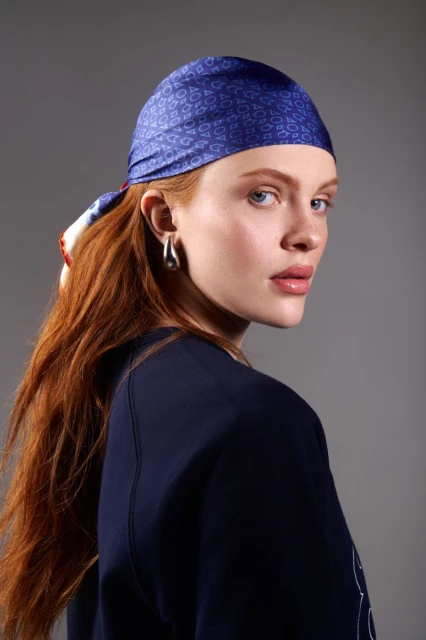 Close-up shot of a fair-skinned woman with red hair and a patterned headscarf looking over her shoulder in front of a gray background.