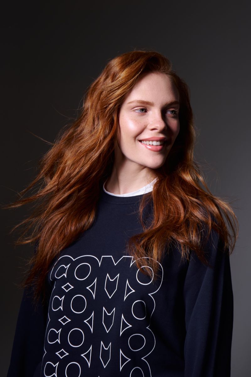 Portrait of a red-haired woman smiling in a blue sweatshirt with white graphic, set against a dark gray background in soft studio lighting.