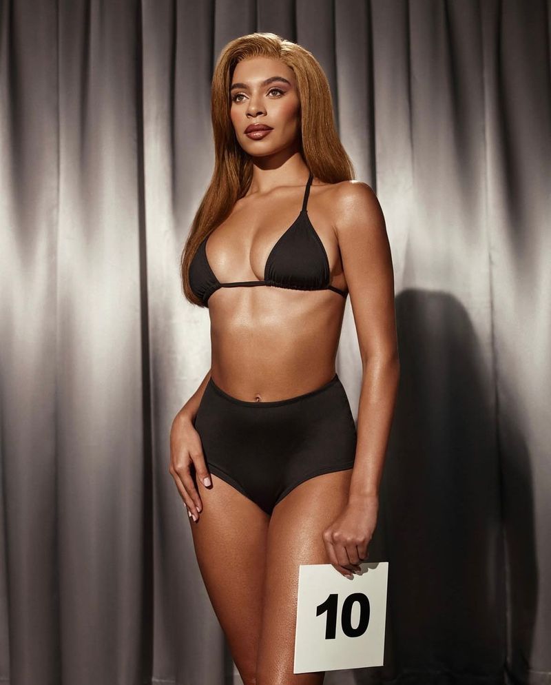 Portrait of a woman in a black bikini against a grey curtain backdrop, holding a card with the number 10. Beauty and fashion focused.