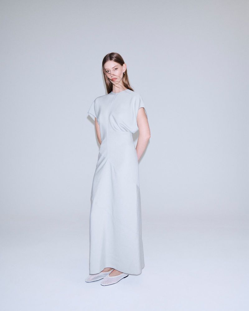 A woman stands in a photo studio wearing a light-blue dress with short sleeves and light-blue shoes against a plain white background.