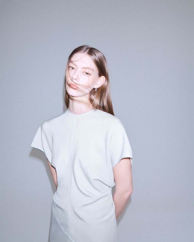 Portrait of a young adult woman with straight brown hair, wearing a light blue dress, posing against a light grey studio backdrop.
