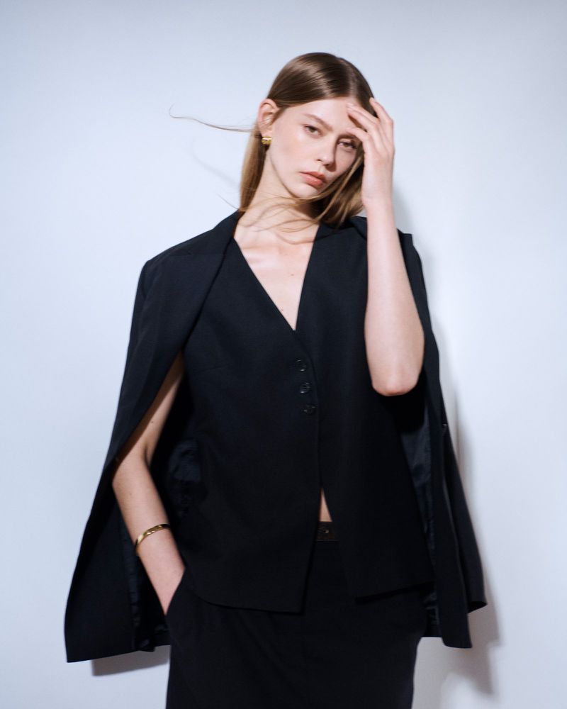 Fashion portrait of a female model with long brown hair in a black blazer-style top with cape sleeves, posing against a white backdrop.