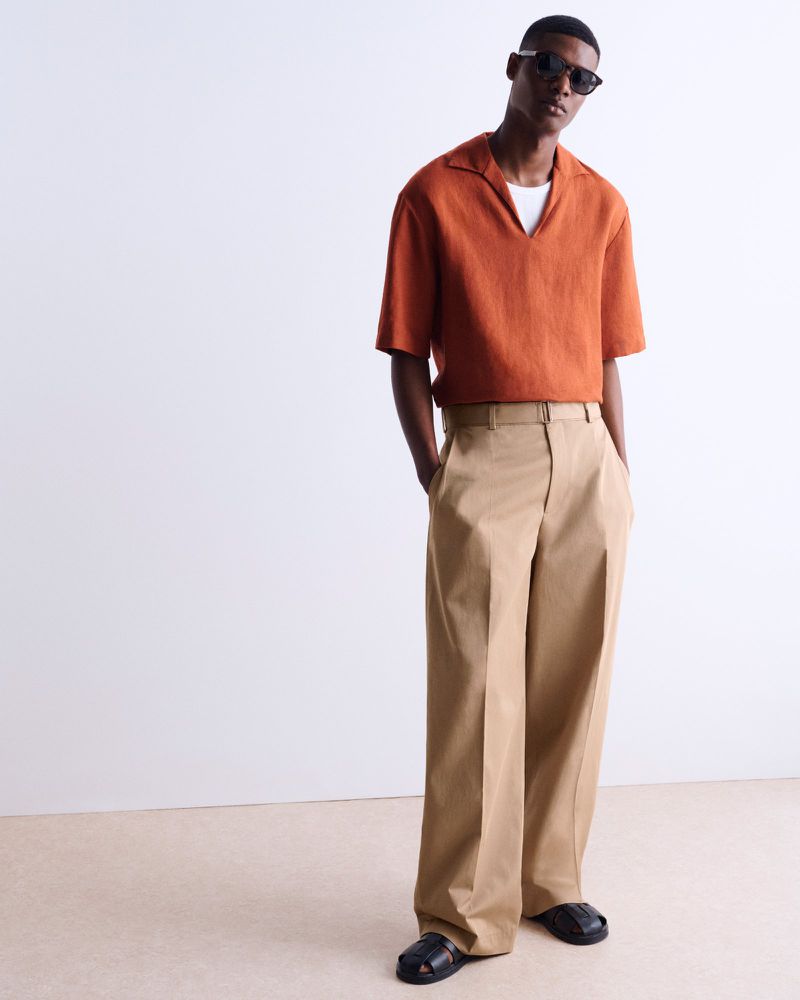 A black male fashion model poses in a burnt-orange short-sleeve shirt and tan wide-legged pants in a minimalist studio setting.