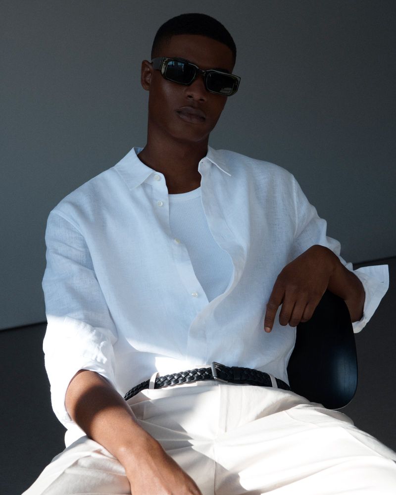 Man in a white shirt and sunglasses poses in front of a gray background. He is seated and looking directly at the camera.