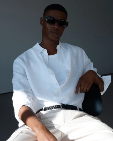 Man in a white shirt and sunglasses poses in front of a gray background. He is seated and looking directly at the camera.
