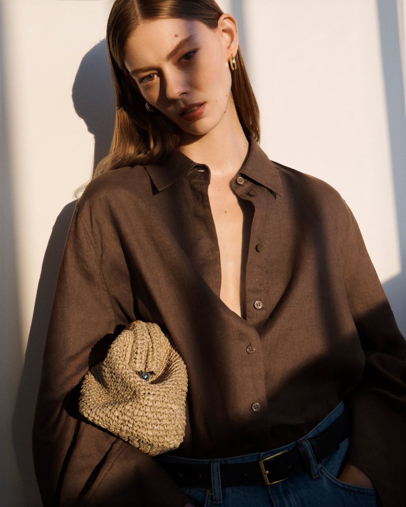 Portrait of a woman wearing a brown button-down shirt and holding a woven handbag indoors with light and shadow on the wall.