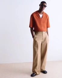 Model wearing dark sunglasses, rust orange polo shirt, and beige trousers against a clean white studio background.