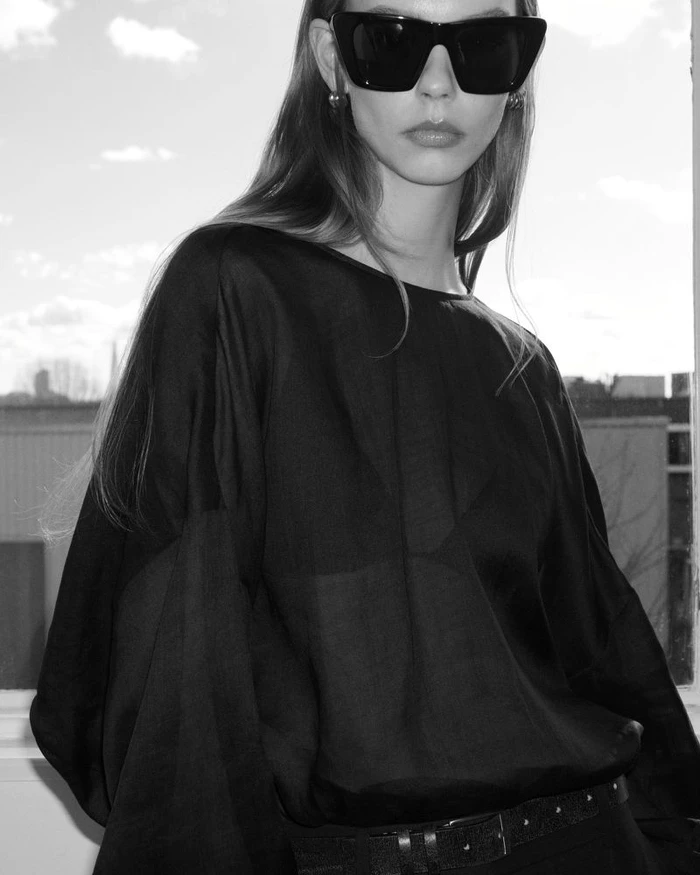Model wearing dark oversized sunglasses and black silk top standing against a window backdrop for COS Linen project.