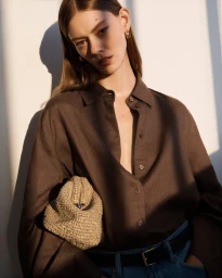 Model wearing a brown button-down shirt holding a woven beige pouch bag against a plain wall.