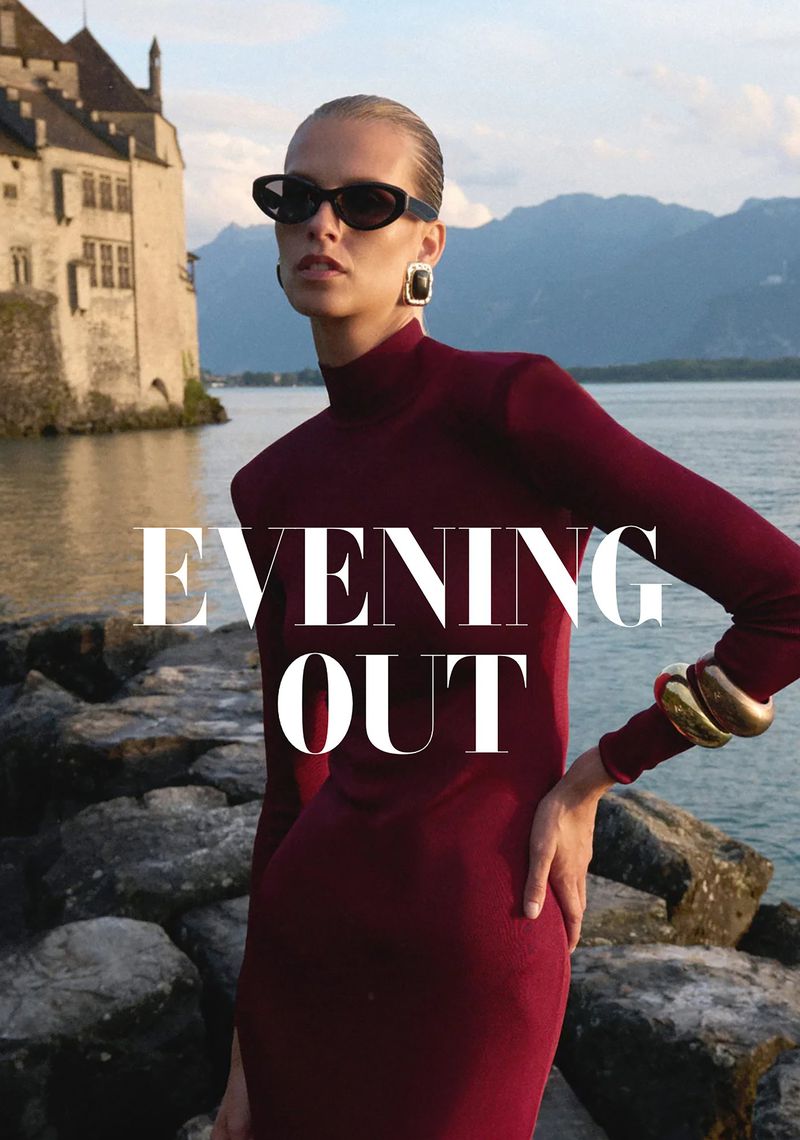 A fair-skinned woman poses in a long-sleeved maroon dress with a lakeside castle and mountains in the background.