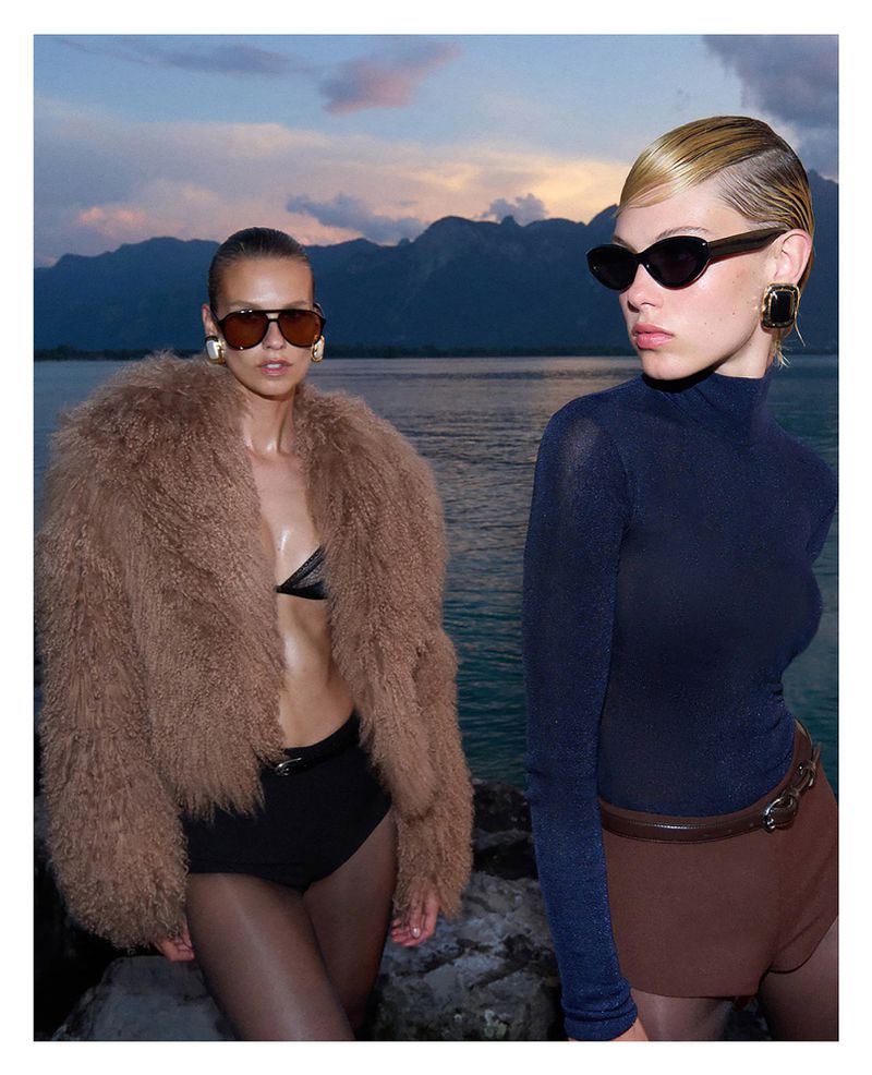Two female models wearing sunglasses and designer winter apparel outdoors by a lake with mountains at dusk.
