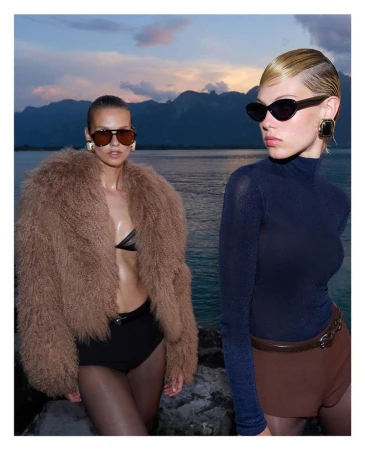 Two female models wearing sunglasses and designer winter apparel outdoors by a lake with mountains at dusk.