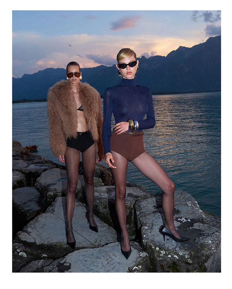 Two women pose on rocky outcrop by a lake at dusk, wearing dark swimsuits, sheer hosiery, heels, and sunglasses with mountain background.