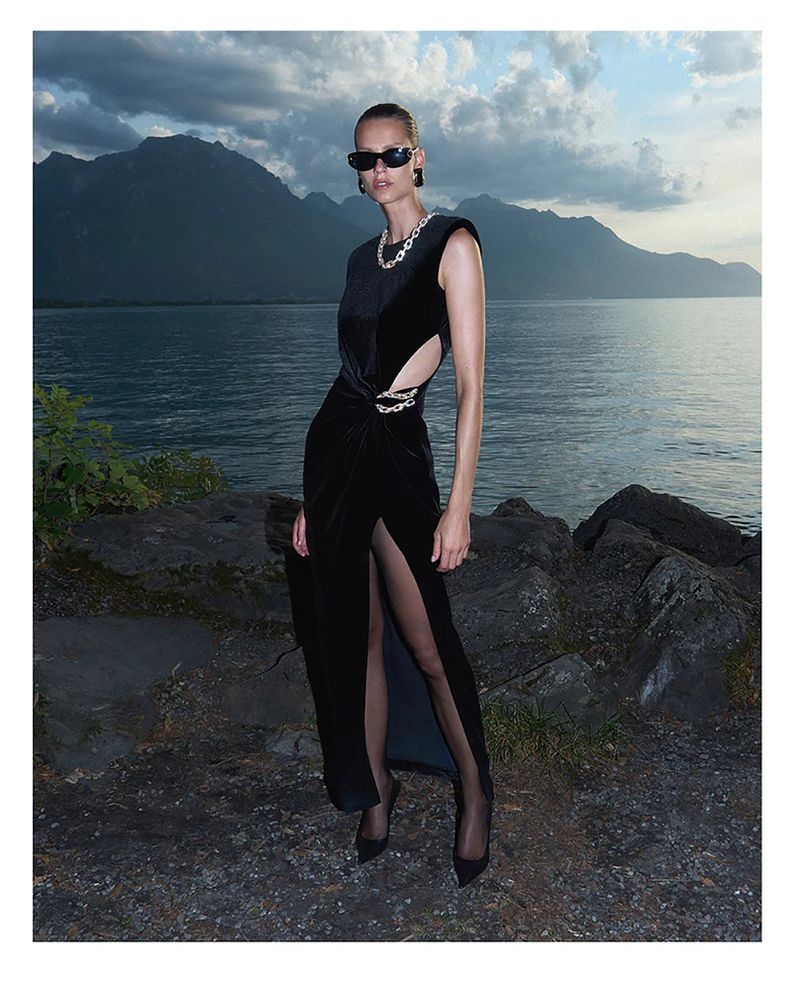 Model wearing a black velvet dress and sunglasses standing by a lake with mountains in the background