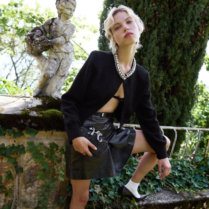 Woman poses with a black studded handbag, short leather skirt, and black jacket in a garden setting with a stone sculpture.