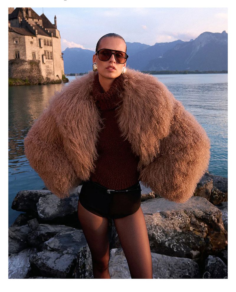 A woman poses in a furry coat on a rocky shore with a castle and lake in the background, illuminated by soft natural light.