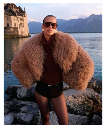 A woman poses in a furry coat on a rocky shore with a castle and lake in the background, illuminated by soft natural light.