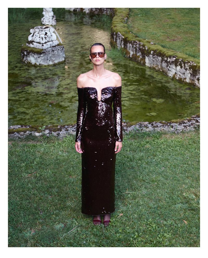 Woman in a long burgundy sequin dress, posing on a lawn near a stone structure with water, wearing dark sunglasses and a neutral expression.