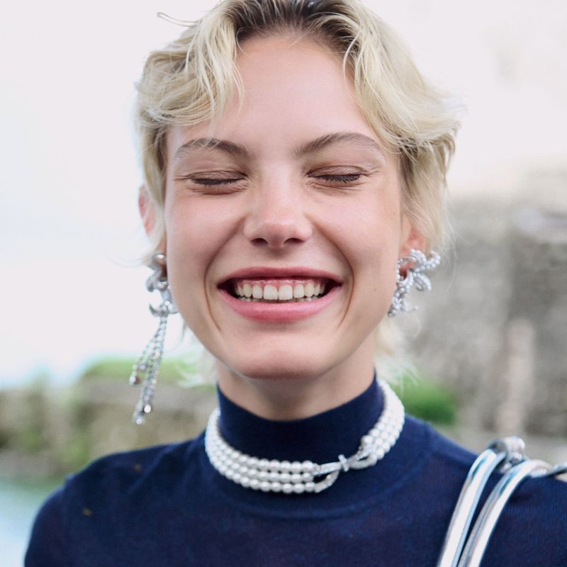 Smiling fair-skinned woman with blonde hair wears pearl choker and sparkling earrings. Close up fashion shot with blurred background.