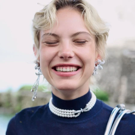 Smiling fair-skinned woman with blonde hair wears pearl choker and sparkling earrings. Close up fashion shot with blurred background.