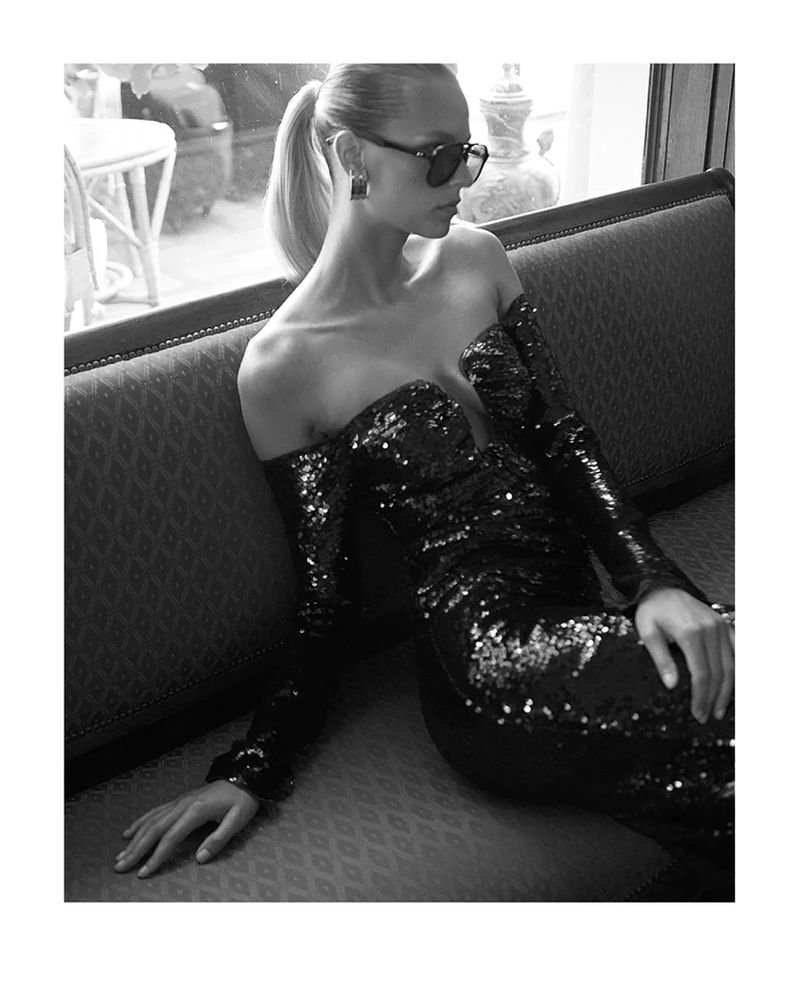 Model in a black sequin off-the-shoulder dress wearing sunglasses seated on a tufted sofa